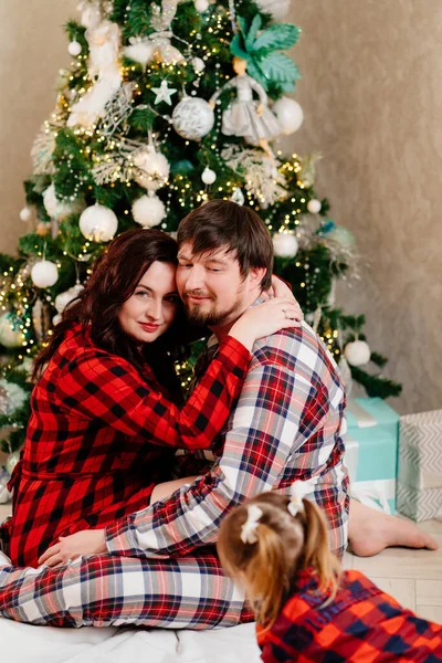Μπαμπά, μαμά και μικρή κόρη με κόκκινες καρό πιτζάμες που παίζουν στο χριστουγεννιάτικο δέντρο. — Φωτογραφία Αρχείου