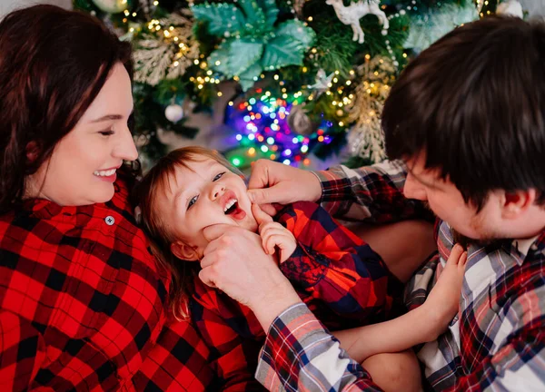 Papá, mamá e hija pequeña en pijama de cuadros rojos jugando en el árbol de Navidad — Foto de Stock