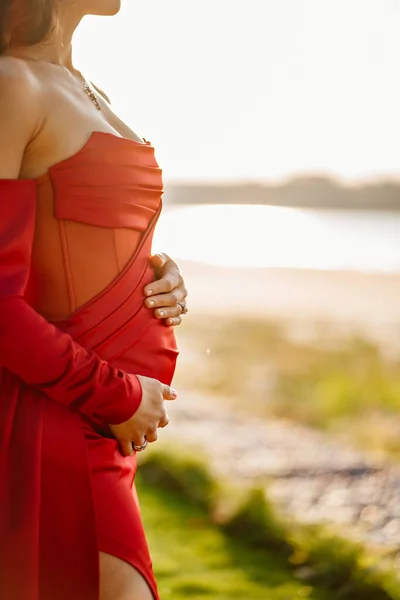Uma mulher grávida em um vestido apertado vermelho acariciando sua barriga com as mãos. — Fotografia de Stock