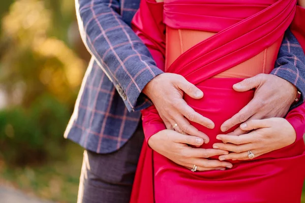 Uomo e donna incinta in un abito rosso stretto accarezzando la pancia con le mani. — Foto Stock