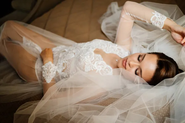 Uma noiva doce em um manto de renda está na cama em um véu. manhã antes do casamento. — Fotografia de Stock