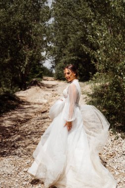 Gelinlikli gelin ağaçlar arasında dağ yolunda yürüyor. Arkadan görünüm. 