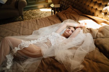 Dantelli bornozlu güzel gelin bir peçenin üzerinde yatakta uzanıyor. Düğünden önce sabah
