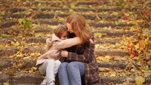 Madre e hija se sientan y se abrazan en las escaleras en el parque de otoño. — Vídeos de Stock