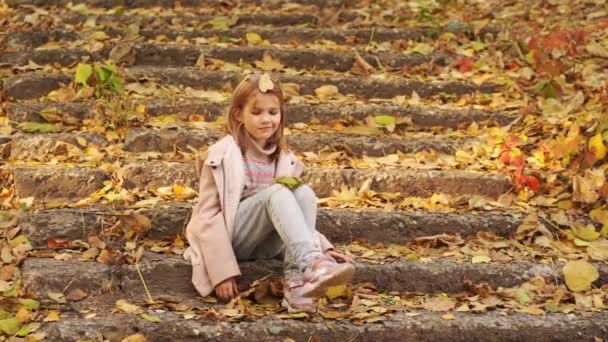 Ein nettes kleines Mädchen sitzt auf einer Treppe mit heruntergefallenem Laub in einem herbstlichen Park. — Stockvideo