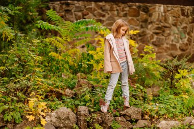 Sonbahar parkında paltolu ve kot pantolonlu, sarı yapraklı şirin bir kız..