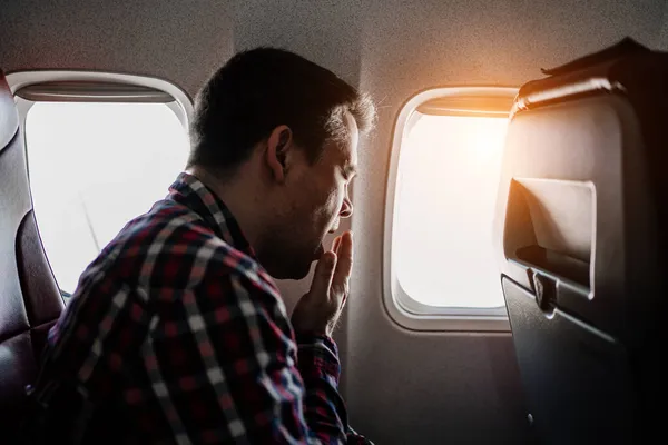 Um homem de camisa xadrez senta-se junto à janela do avião e boceja. voo longo. — Fotografia de Stock