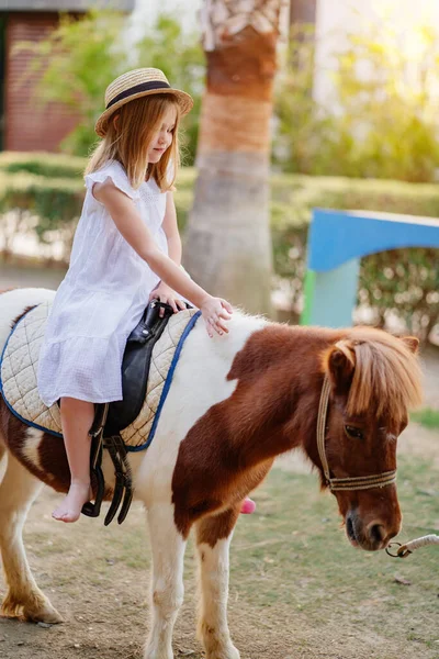 Une petite fille en robe blanche et chapeau de paille assis sur un poney. — Photo