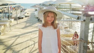 Beyaz elbiseli ve geniş kenarlı şapkalı küçük bir kız iskelede duruyor.