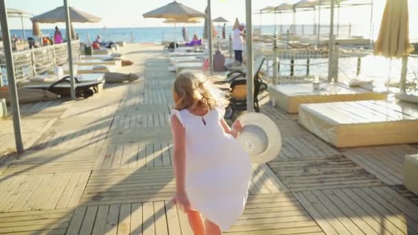 Маленька дівчинка блондинка в білій сукні і широкий капелюх біжить на пірсі — стокове відео