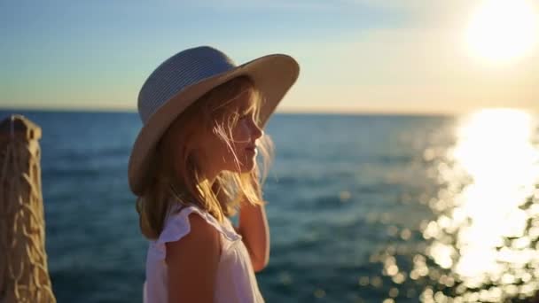 Una niña rubia con un vestido blanco y un sombrero de ala ancha está junto al mar — Vídeo de stock