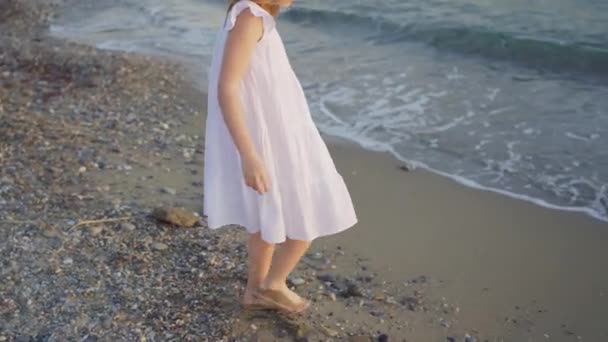 Una linda niña en un vestido blanco camina descalza en el agua fría del mar. — Vídeos de Stock