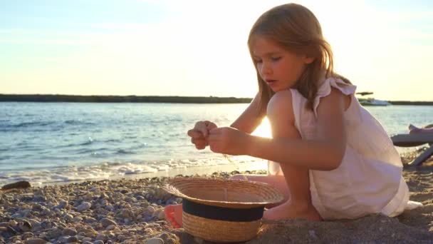 Fille en robe blanche se trouve sur le bord de la mer et recueille des cailloux et des brindilles en chapeau de paille — Video