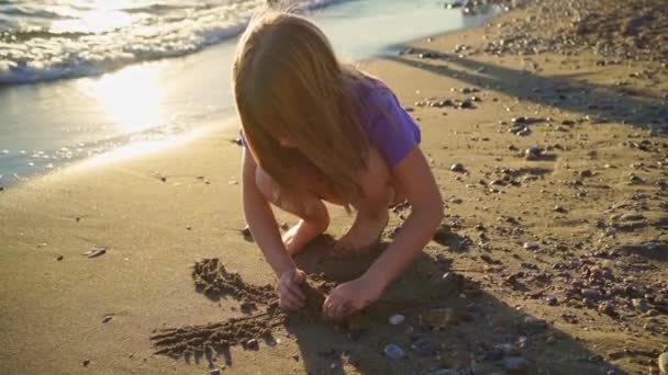 Una linda niña se sienta en la orilla del mar, jugando en la arena y con guijarros. — Vídeos de Stock