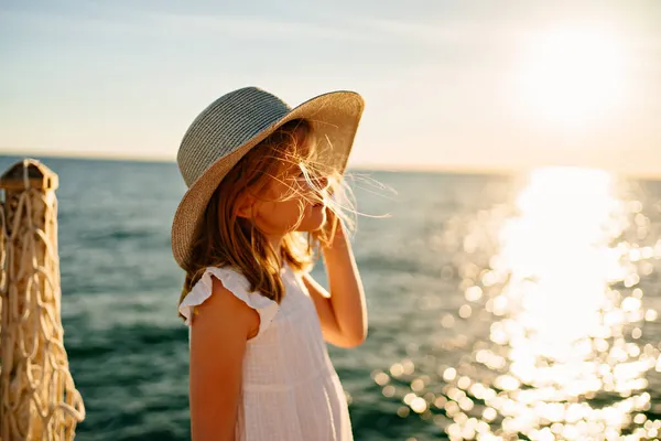 Маленькая блондинка в белом платье и широкополой шляпе стоит у моря — стоковое фото
