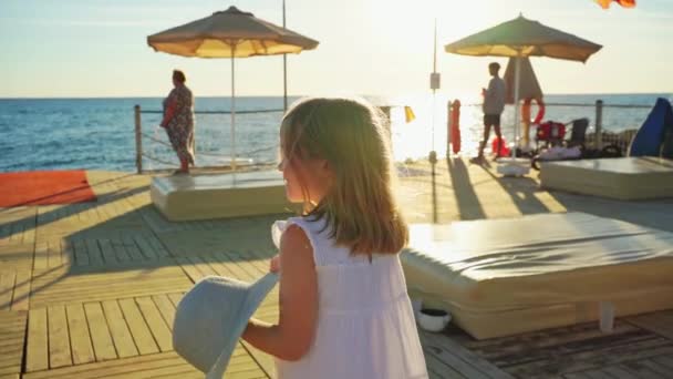 Een meisje blond in een witte jurk met een brede hoed loopt op de pier — Stockvideo