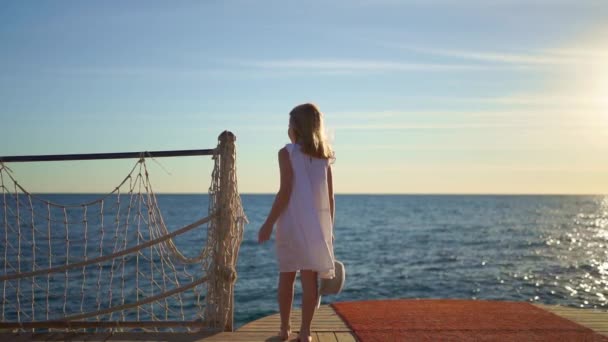 İskeledeki denizin kenarında geniş kenarlı şapkalı beyaz elbiseli küçük bir kız. — Stok video