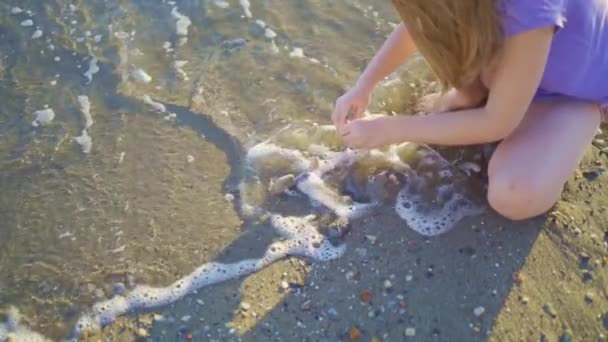 Una linda niña se sienta en la orilla del mar, jugando en la arena y con guijarros. — Vídeos de Stock