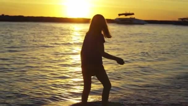 At sunset little girl walks along seashore and throws stones into water. — Stock Video