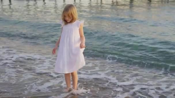 Beyaz elbiseli şirin küçük bir kız soğuk deniz suyunda yalınayak yürüyor.. — Stok video