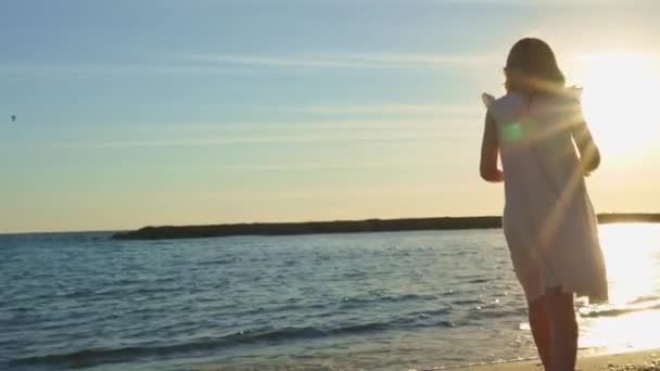 Девушка в белом платье на берегу моря выливает гальку и ветки из соломенной шляпы — стоковое видео