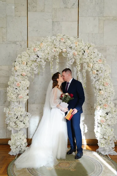 Νύφη και γαμπρός φιλιούνται σε μια αψίδα λουλουδιών με πιστοποιητικό γάμου. — Φωτογραφία Αρχείου