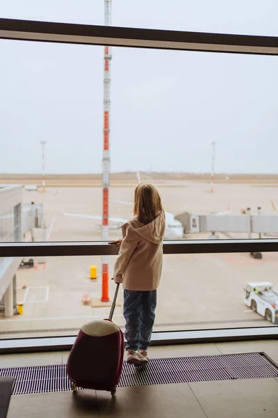 Mała dziewczynka z walizką stoi przy oknie na lotnisku. — Zdjęcie stockowe