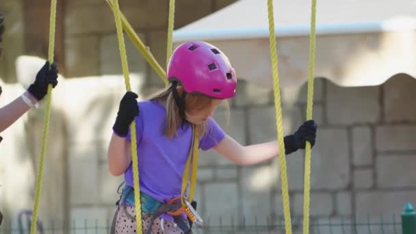 Una niña valiente en un casco, camiseta púrpura y pantalones cortos sube en un parque de cuerdas — Vídeo de stock