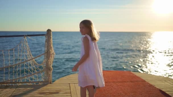 Een klein meisje in een witte jurk met een brede hoed aan zee op de pier — Stockvideo