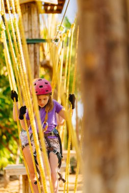 Miğferli kız, tişört ve şortla halat parkına tırmanıyor.
