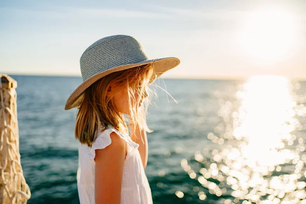 Маленькая блондинка в белом платье и широкополой шляпе стоит у моря — стоковое фото