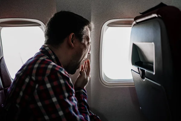Un hombre con una camisa a cuadros se sienta junto a la ventana del avión y bosteza. vuelo largo. — Foto de Stock