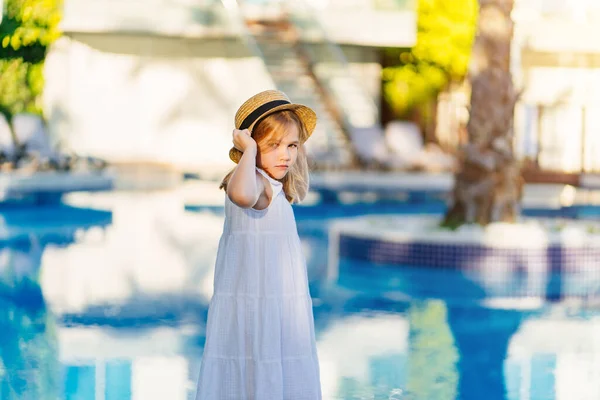 Linda niña en un vestido blanco y sombrero de paja junto a la piscina cerca de la villa. — Foto de Stock