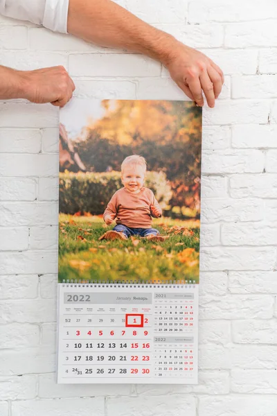 Las manos de los hombres sostienen el calendario con la foto del niño delante de la pared de ladrillo blanco — Foto de Stock