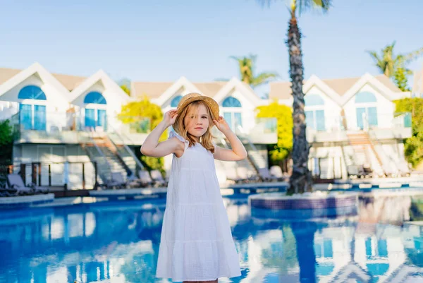 Linda niña en un vestido blanco y sombrero de paja junto a la piscina cerca de la villa. — Foto de Stock