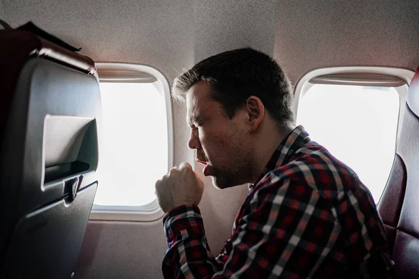 Um homem com uma camisa xadrez tosse no avião em um assento da janela. — Fotografia de Stock