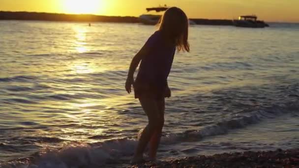 На заході сонця маленька дівчинка гуляє уздовж узбережжя моря і кидає каміння у воду . — стокове відео