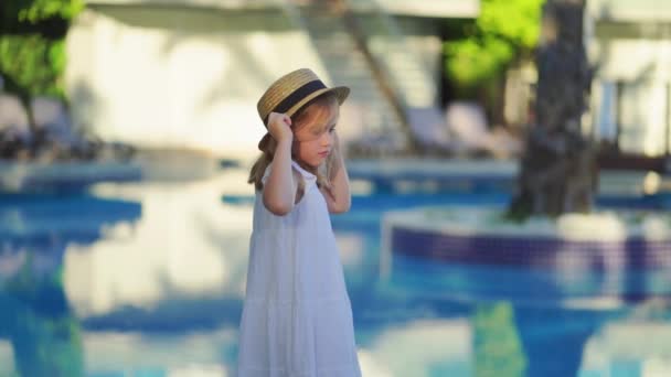 Cute little girl in a white dress and straw hat by the pool near the villa. — Stock Video