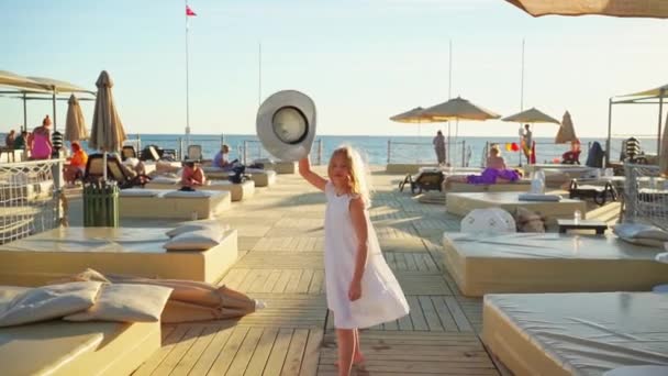 Een meisje blond in een witte jurk met een brede hoed loopt op de pier — Stockvideo