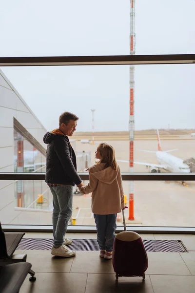 Папа и дочь с чемоданом стоят у окна аэропорта. — стоковое фото