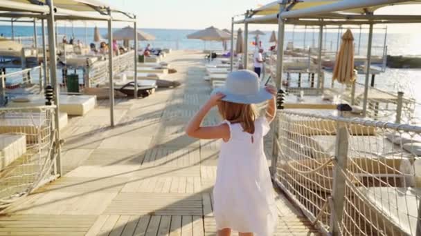 Una niña rubia con un vestido blanco y un sombrero de ala ancha corre por el muelle — Vídeos de Stock