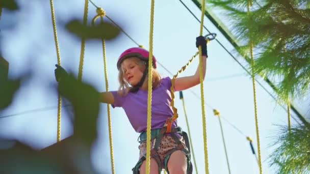 Una valiente niña en un casco, camiseta púrpura y pantalones cortos sube a un parque de cuerdas — Vídeo de stock