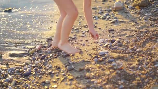 Al atardecer. piernas de niña caminando a lo largo de la orilla del mar y arrojando piedras al agua — Vídeos de Stock