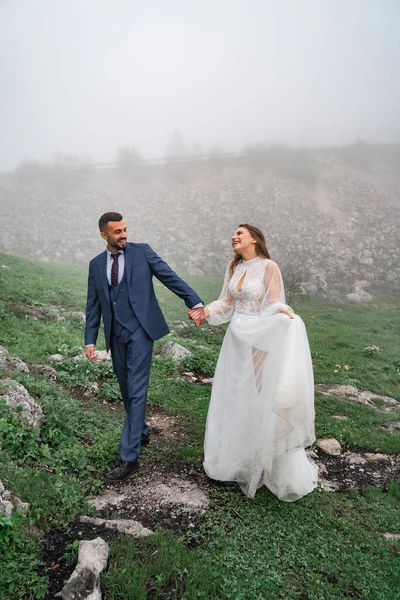 新婚夫婦は手を取り合って霧の中を歩く。旅行中の結婚式 — ストック写真