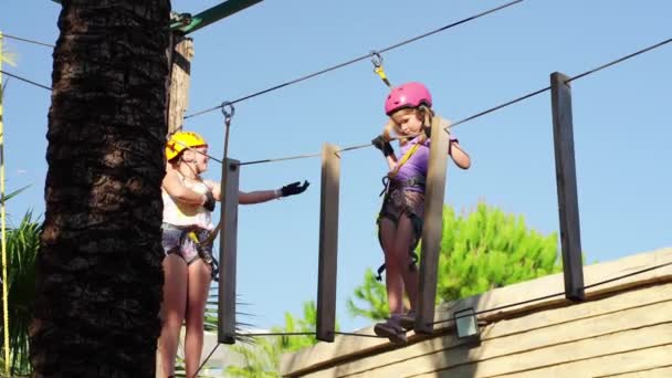 Dos chicas asustadas pero valientes en casco, camiseta y pantalones cortos sube en el parque de cuerdas — Vídeo de stock