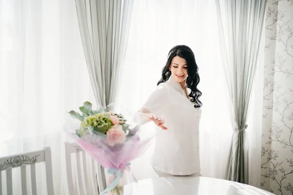 Novia embarazada morena en pijama blanco por ventana y mesa con ramo. — Foto de Stock