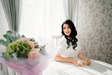 Beyaz ipek pijamalı, makyajlı gelin masada çay içiyor.