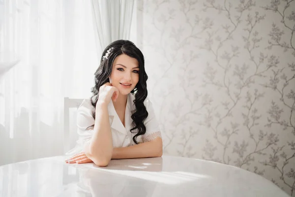 Brunette bride in white silk pajamas with styling and makeup at the table — Stockfoto
