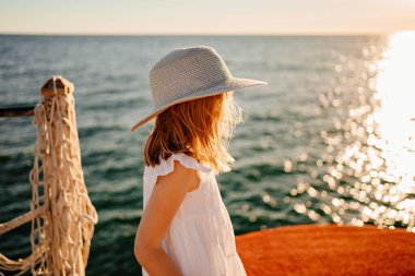 a little girl blonde in a white dress and a wide-brimmed hat stands by the sea  clipart