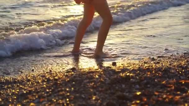 Ao pôr-do-sol. pernas de menina andando ao longo da costa e jogando pedras na água — Vídeo de Stock